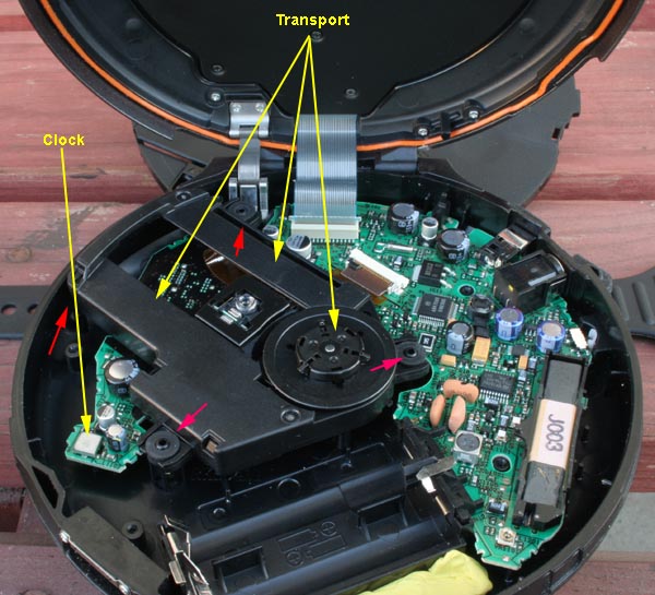 Here's what you will see next. Note the the position of the clock (aka the oscillator). Delicately cover it and the surrounding capacitors with a little bit of rope caulk or adhesive tacky putty. Careful: do not get any caulk/tack on the transport or the four transport-suspension struts (red arrows). 